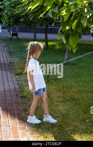 Little girl walking under the water from the sprinkler irrigation in the summer city park. Childhood, leisure and people concept - happy child rest an Stock Photo