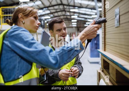 Young man with Down syndrome working in warehouse, female colleague teaching him scan products with scanner. Concept of workers with disabilities Stock Photo