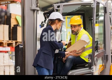 Forklift driver talking with manager in modern industrial factory. Warehouseman reading order, order picking. Warehouse worker preparing products for Stock Photo