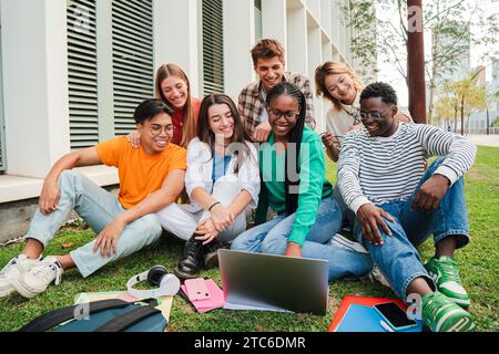 Portrait of multiracial college students using a laptop computer while having fun watching funny movie sitting on lawn after doing homework outside university campus. Smiling friends taking a break. High quality photo Stock Photo