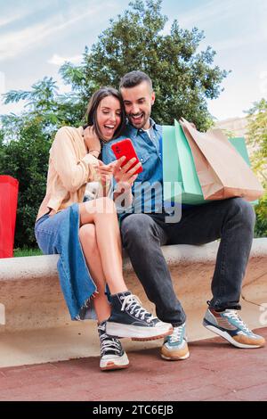 Vertical. Surprised couple using a cellphone to find sales, shopping online sitting with bags. Man and woman having fun with a smartphone app. Young real people browsing with a mobile phone at mall. High quality photo Stock Photo