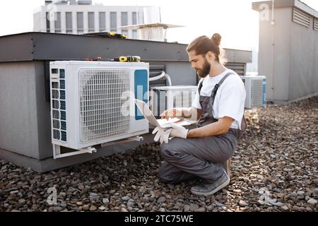 Repairman looking at conditioner and using laptop on roof Stock Photo