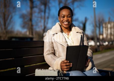 Outdoor portrait of happy african-american woman on sunny day. She is sitting on bench in the street and using digital tablet. Stock Photo