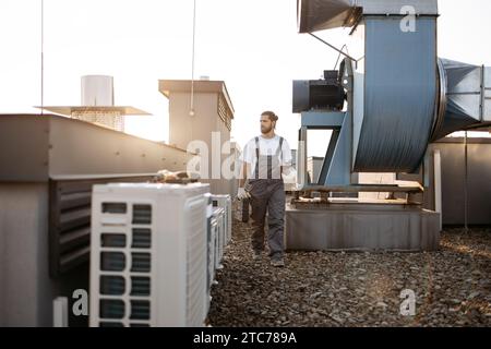Concentrated bearded man wearing uniform holding toolkit and using digital tablet on factory. Caucasian professional craftsman walking by air conditio Stock Photo
