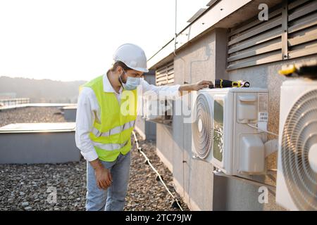 Adult qualified craftsman wearing mask to avoid breathing dust while inspecting air conditioner outdoors. Focused male in helmet and vest installing d Stock Photo