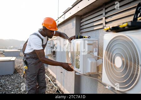 African american factory worker in orange hard hat standing and holding hands on air conditioner while checking attachment to wall outdoors. Concept o Stock Photo