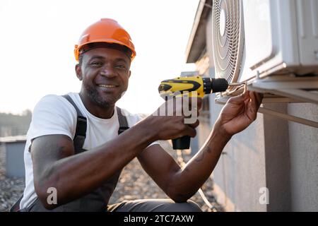 Portrait of handsome african american male using electrical screwdriver for repairing ventilation system outdoors. Smiling man with protective helmet Stock Photo