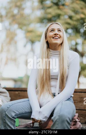 Gorgeous, blonde, female student having school break. She is sitting on a bench, having fun conversation with her classmates and smiling. Stock Photo