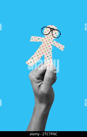 the hand of a man in black and white holding a paper man doll, wearing eyeglasses, on a blue background, as a prank for the innocents day, a feast hel Stock Photo