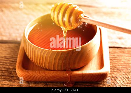 Natural honey dripping from dipper into wooden bowl on table under sunlight, closeup Stock Photo