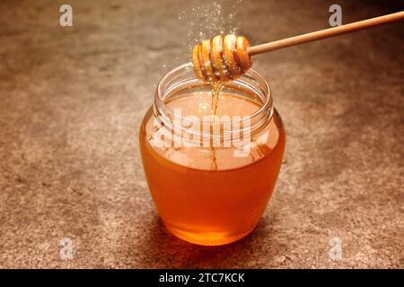 Natural honey dripping from dipper into glass jar on table under sunlight Stock Photo