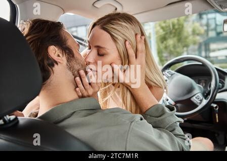 charming blonde woman kissing man on drivers seat in modern car on city street, urban romance Stock Photo