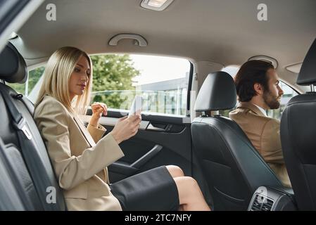 blonde elegant businesswoman using smartphone near elegant man driving luxury car on street Stock Photo