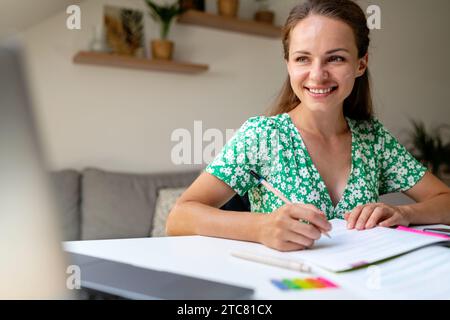 Adult student e-learning. Attractive brunette woman Smiling while Using Workbook at home desk. Educational Elegance. Stock Photo