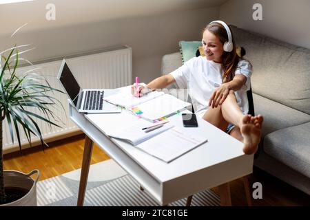 Home education. Attractive brunette woman in headphones using workbook at home desk. Using laptop for e-learning. Stock Photo