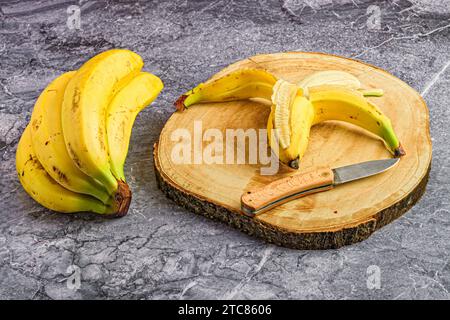 A ripe, yellow banana sits partially peeled on a wooden board, alongside a bunch of unpeeled bananas Stock Photo