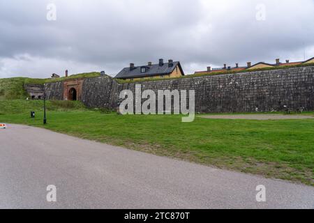 A countryside road with a brick wall running alongside it, set amongst a lush green landscape with grass growing atop the wall Stock Photo