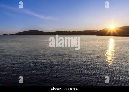 A summer evening's midnight sun peeks over a hill, casting golden rays on a fjord near port of Bodo, under a clear blue sky in Norway's Lofoten Island Stock Photo