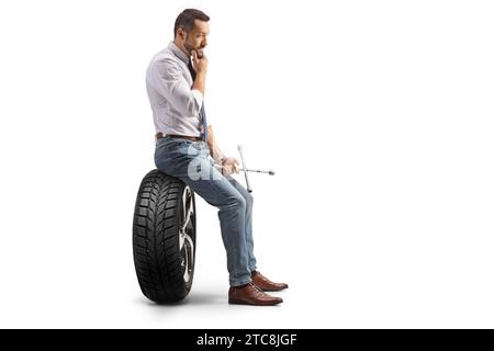 Profile shot of a pensive man sitting on a car tire and holding a lug wrench isolated on white background Stock Photo