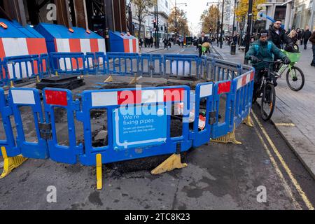 Cyclists mount the pavement where Thames Water utility barriers surround street works where they are fixing pipes on a cordoned off Oxford Street on 6th December 2023 in London, United Kingdom. Thames Water Utilities Ltd is a large private utility company responsible for the water supply and waste water treatment in most of Greater London and surrounding areas in England. Stock Photo