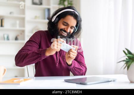 Cheerful Indian Man In Wireless Headphones Playing Video Games On Smartphone Stock Photo