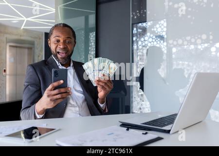 Shocked and happy african american male businessman sitting in office holding money fan and looking surprised at phone screen. Stock Photo