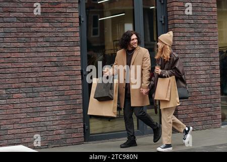 Happy young couple in stylish casualwear walking along wall of modern building after shopping and having coffee Stock Photo