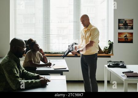 Mature male coach with carbine or automatic rifle standing in front of African American students and explaining them how to shoot Stock Photo