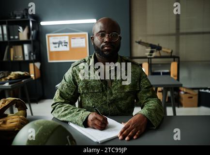 Young serious African American man in camouflage uniform sitting by desk, making notes and listening to explanation of teacher Stock Photo