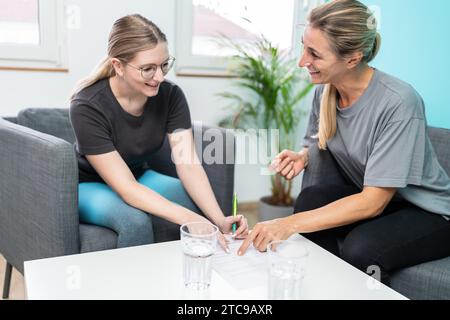 Two women smiling and engaging in conversation with one writing on a form in a well-lit therapy room Stock Photo