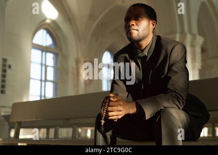 Portrait of young Black man as priest holding rosary beads in church and praying in sunlight rays, copy space Stock Photo