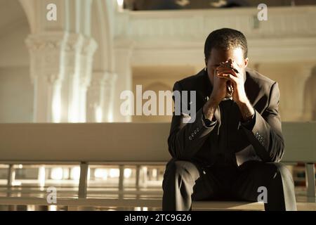 Front view at young African American priest praying in church and holding rosary beads in sunlight, copy space Stock Photo