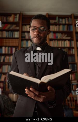 Vertical portrait of young African American man as priest reading Bible in church with books in background Stock Photo