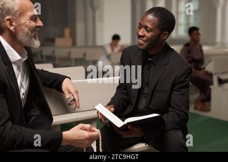 Portrait of young Black man as smiling priest talking to senior man after Sunday service in catholic church, copy space Stock Photo