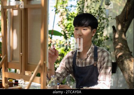 A positive and handsome young Asian man in an apron is creating his artwork, painting with water color on a canvas easel in his home garden. Leisure a Stock Photo