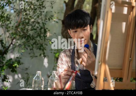 A positive and handsome young Asian man in an apron is creating his artwork, painting with water color on a canvas easel in his home garden. Leisure a Stock Photo