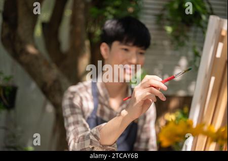 Young positive and handsome Asian man is enjoying painting on a canvas on an easel in his home garden on a bright day. Home leisure, creative activity Stock Photo
