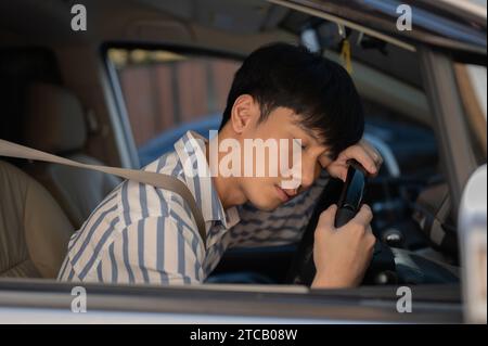 A tired young-adult Asian man is falling asleep in his car after driving for long hours, taking a break. People and vehicle concepts Stock Photo