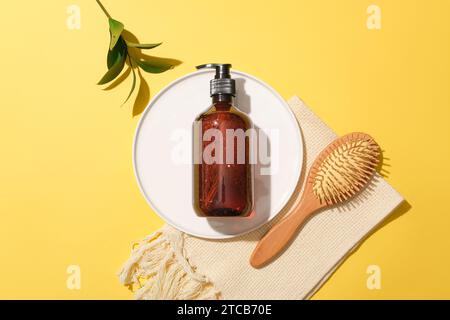 View from above of a brown shampoo bottle on a white ceramic plate, a detangling comb, green leaves and a towel on a yellow background. Cosmetic mocku Stock Photo