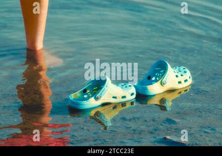 Rubber slippers float in clear water next to female legs closeup Stock Photo