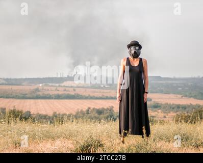 Young girl in a black dress and gas mask on the background of smoking factory chimneys Stock Photo
