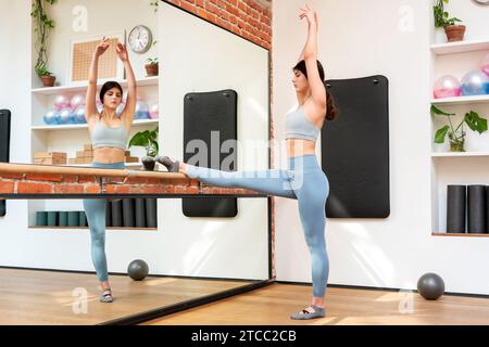 Full body side view of young ballerina in activewear doing stretching exercises with raised arms at barre in front of mirror in gym Stock Photo