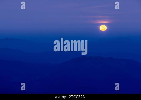 The full moon shines over blue mountains at dusk, the full moon against a dark blue sky in the background. Stock Photo