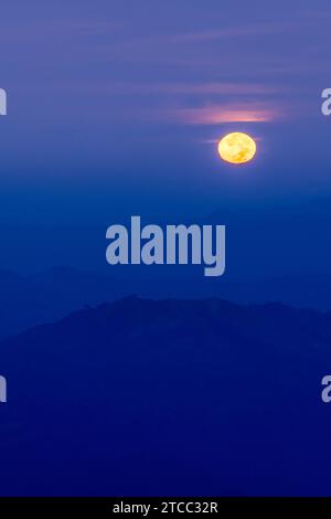 The full moon shines over blue mountains at dusk, the full moon against a dark blue sky in the background. Stock Photo