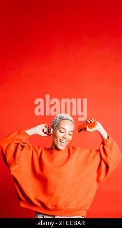 Happy young woman dancing with arms raised while wearing wireless earphones in a studio. Woman listening her favourite music playlists. Stock Photo