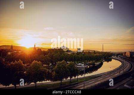 A view of a bustling urban landscape, featuring several cars driving down a road which leads to a stunning sunset over the water Stock Photo