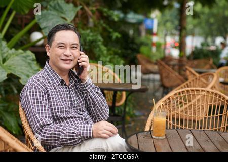 Portrait of smiling senior man sitting at cafe table, drinking ice coffee and talking on phone Stock Photo