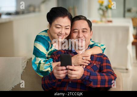 Smiling senior woman hugging husband from behind when he is taking selfie Stock Photo