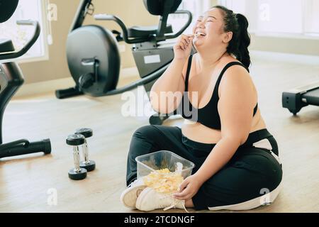 Binge Eating Disorder (BED), happy fat women laugh enjoy unstopable eating chips snack food during relax brake from exercise in fitness sport club. Stock Photo