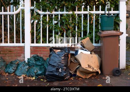 A brown wheelie bin, cardboard, and black bin bags are on the pavement waiting for refuse collection. Outside and house with a white painted fence. Stock Photo